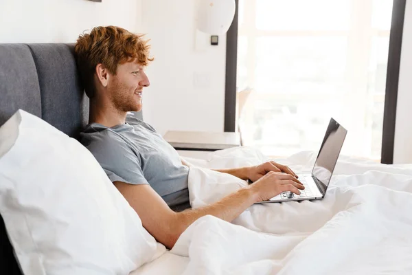 Genç Gülümseyen Gri Tişörtlü Laptopla Çalışan Evde Yatağında Yatan Kızıl — Stok fotoğraf
