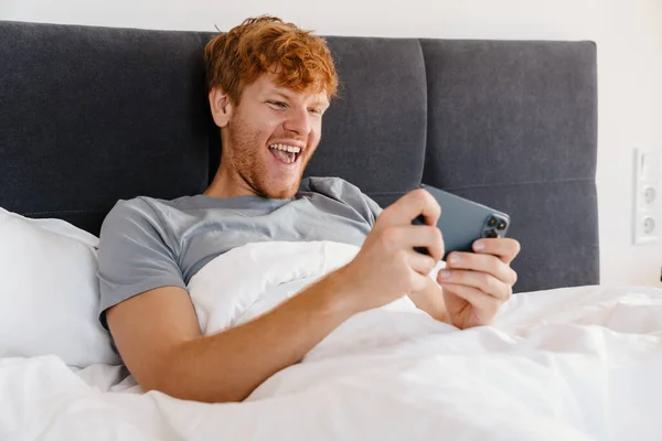 Young handsome enthusiastic redhead man in gray t-shirt playing mobile game on his phone, while lying on bed at home