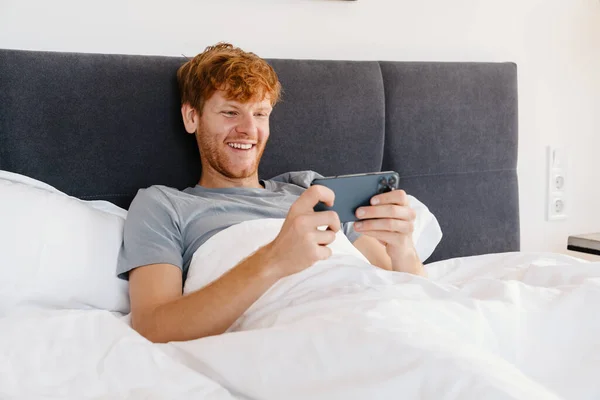 Young handsome smiling redhead man in gray t-shirt holding and using his phone, while lying on bed at home