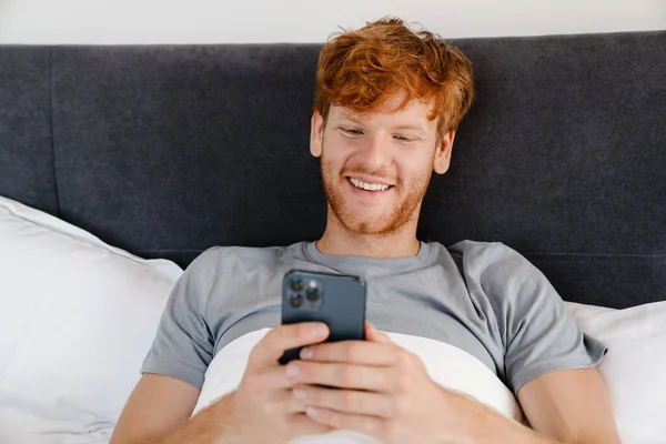 Gri Tişörtlü Telefonuyla Evde Yatarken Gülümseyen Kızıl Saçlı Genç Adam — Stok fotoğraf