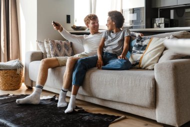 Genç, yakışıklı, gülümseyen adam, güzel Afrikalı kız arkadaşına bakıyor ve uzaktan kumandayla TV kanalını değiştiriyor.