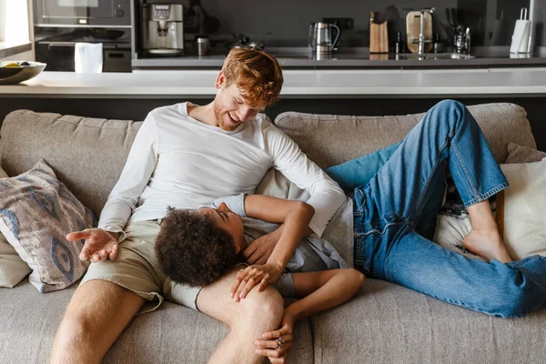 stock image Young handsome smiling redhead guy caressing his beautiful african girlfriend liying on his knee , while they are talking on couch at home