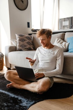 Genç kızıl saçlı adam evde otururken gülümsüyor ve dizüstü bilgisayar kullanıyor.