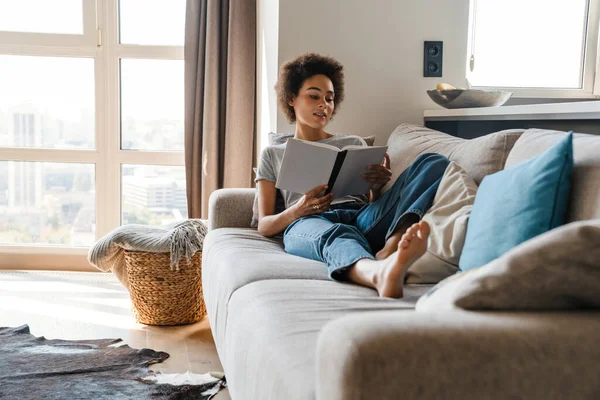 年轻的卷曲女人穿着牛仔裤看书 躺在家里的沙发上 — 图库照片