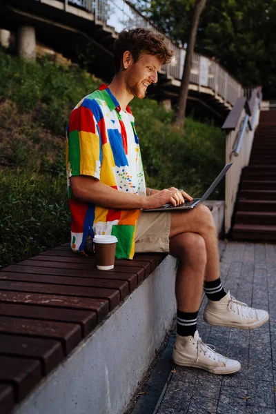 公園のベンチにコーヒーと一緒に座っている間 ノートパソコンで作業している若いハンサムな笑顔の赤毛の男 — ストック写真