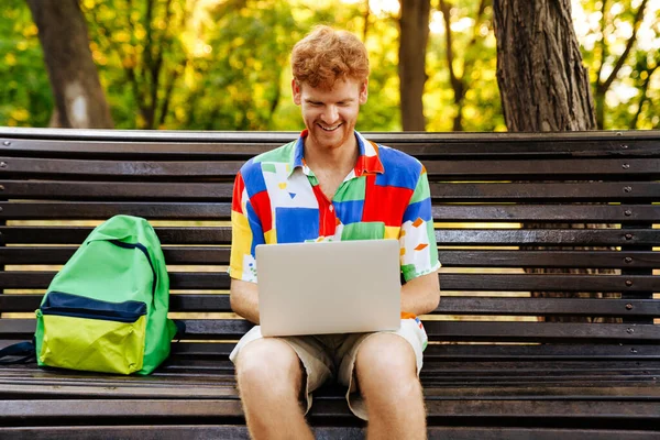 Mladý Pohledný Usměvavý Zrzka Muž Barevné Košili Pracuje Notebookem Zatímco — Stock fotografie
