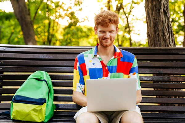 公園のベンチにバックパックで座っている間 ラップトップで作業カラフルなシャツの若いハンサムな笑顔の赤毛の男 — ストック写真