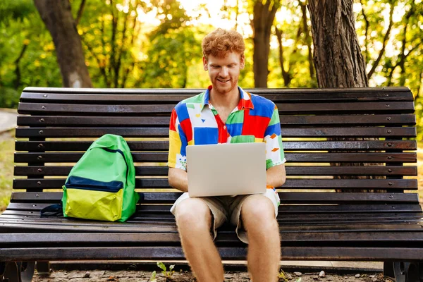 Młody Przystojny Uśmiechnięty Rudy Mężczyzna Kolorowej Koszuli Pracujący Laptopem Siedzący — Zdjęcie stockowe