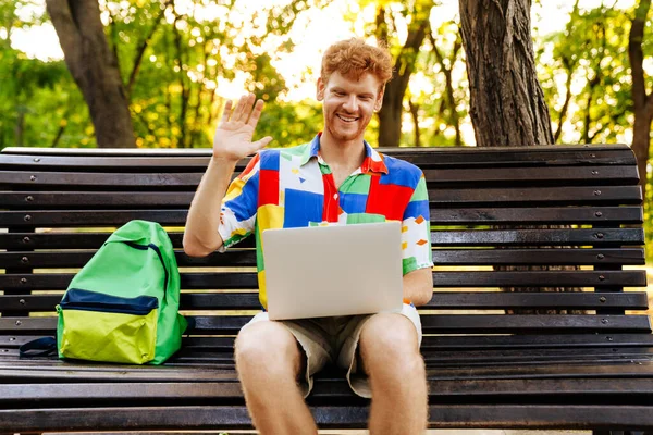 Młody Przystojny Uśmiechnięty Rudzielec Laptopem Wideokonferencji Machający Plecakiem Ławce Parku — Zdjęcie stockowe