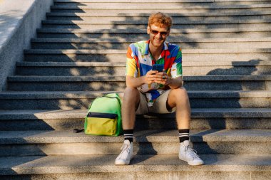 Dışarda sırt çantasıyla otururken renkli gömlekli, güneş gözlüklü ve telefonunu kullanan yakışıklı bir adamın portresi.