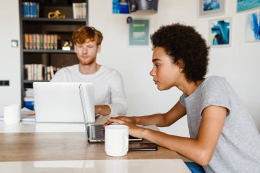 Genç, sakin, kızıl saçlı bir adam ve güzel bir Afrikalı kadın dizüstü bilgisayarlarıyla evde oturuyor.