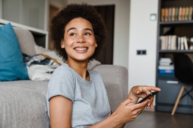Güzel, gülümseyen kıvırcık Afrikalı bir kadının portresi. Elinde telefon, yana bakıyor. Evdeki kanepede uzanıyor.