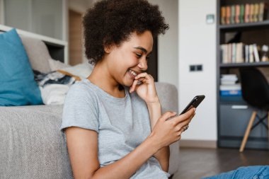 Güzel, gülümseyen kıvırcık Afrikalı bir kadının portresi. Telefonu tutuyor, kullanıyor ve dudaklarına dokunuyor. Evdeki kanepede uzanıyor.
