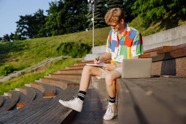 Młody Przystojny Stylowy Skupiony Mężczyzna Okularach Robi Notatki Siedząc Laptopem — Zdjęcie stockowe
