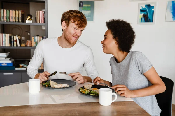 stock image Young beautiful smiling interracial couple looking at each other during breakfast. Young handsome redhead guy and beautiful african curly woman at home