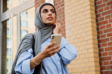 Düşünceli orta yaşlı Müslüman kadın tesettür takıyor. Şehir caddesinde dikilirken akıllı telefon kullanıyor.