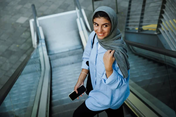 年轻的穆斯林妇女站在室外自动扶梯上用手机戴头巾 — 图库照片