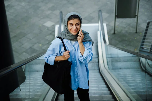 屋外のエスカレーターに立っている間 携帯電話を使用してスカーフを身に着けている若いイスラム教徒の女性 — ストック写真
