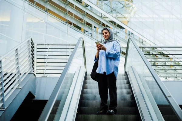屋外のエスカレーターに立っている間 携帯電話を使用してスカーフを身に着けている若いイスラム教徒の女性 — ストック写真