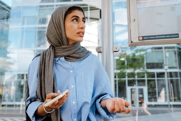 年轻的穆斯林妇女坐在巴士站外 头戴头巾 用手机 — 图库照片