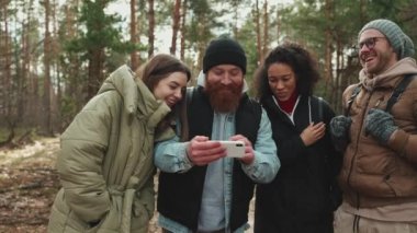 Çok uluslu turist arkadaşları ormanda telefonla video izlerken gülüyorlar.