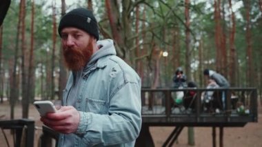 Gülümseyen sakallı turist cep telefonundan mesaj atıyor ve ormanda kamp ateşinin etrafında arkadaşlarıyla geziyor.