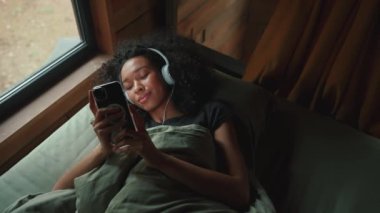 Ormandaki bir modüler evde müzik dinleyen ve cep telefonuyla mesajlaşan pozitif Afrikalı kadın.