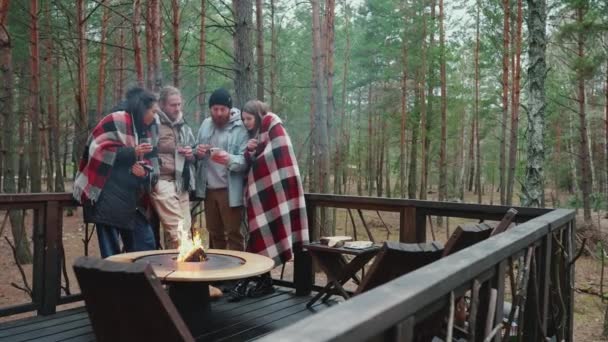 森の中の毛布で覆われたキャンプファイヤーの近くで電話をしながら話している多国籍の友人を笑って — ストック動画