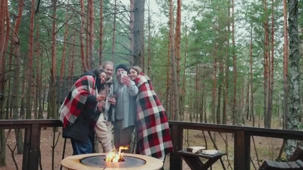 森の中の毛布で覆われたキャンプファイヤーの近くの電話でビデオ通話で話す積極的な多国籍の友人 — ストック動画