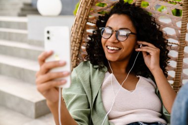 Heyecanlı, gülümseyen, kulaklıklı Afro-Amerikan kadın, dışarıda sandalyede otururken akıllı telefon kullanıyor.
