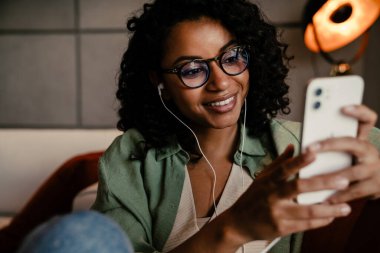 Evde otururken kulaklıklı neşeli, Afro-Amerikalı kadın akıllı telefon kullanıyor.