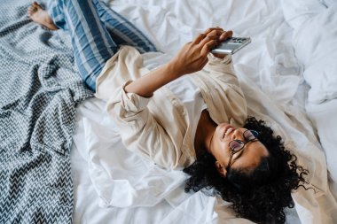 Gözlüklü siyahi bir kadın evde yatağında dinlenirken cep telefonu kullanıyor.