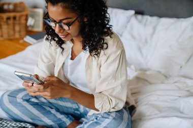 Gözlüklü siyahi bir kadın evde yatağında dinlenirken cep telefonu kullanıyor.