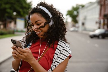 Dışarıda yürürken kulaklık takan siyah bir kadın cep telefonu kullanıyor.