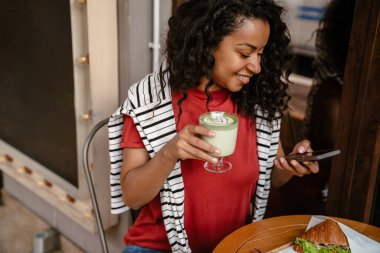 Genç siyahi kadın cep telefonu kullanıyor ve açık kafede öğle yemeği yiyor.