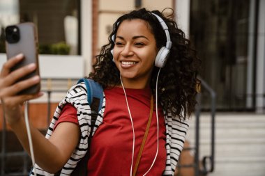 Kulaklıklı genç siyahi kadın dışarıda beklerken cep telefonu kullanıyor.