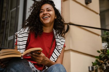 Genç siyahi kadın, merdivenlerde oturmuş kitap okuyor ve gülümsüyor.