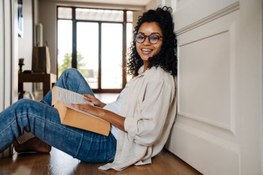 Afro kıvırcık saçlı Afro-Amerikalı bir kadın evde oturmuş kitap okuyor.