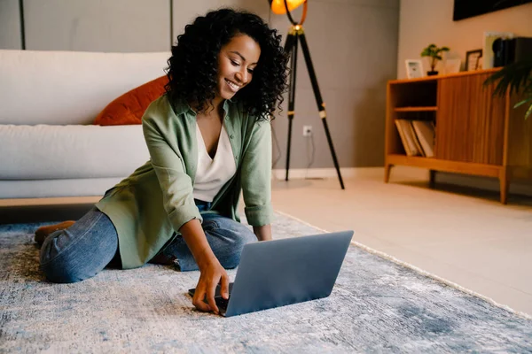 快乐的年轻非洲女人坐在家里的地板上 一边用笔记本电脑 — 图库照片