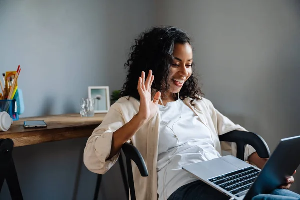 年轻黑人妇女坐在家里的椅子上 一边挥手一边用笔记本电脑工作 — 图库照片