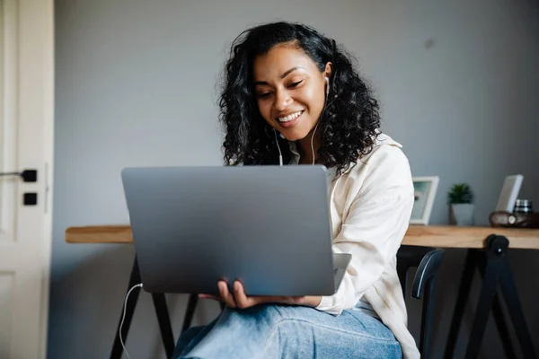 年轻黑人妇女坐在家里的椅子上 一边笑一边用笔记本电脑工作 — 图库照片