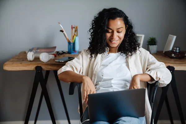 年轻黑人妇女坐在家里的椅子上 一边笑一边用笔记本电脑工作 — 图库照片