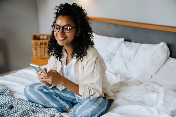 Gözlüklü Siyahi Bir Kadın Evde Yatağında Dinlenirken Cep Telefonu Kullanıyor — Stok fotoğraf