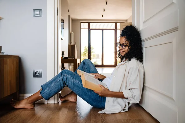 年轻的黑人女子坐在家里看书时戴着眼镜 — 图库照片