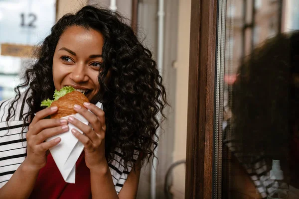Nuori Musta Nainen Hymyilee Syö Voileipää Istuessaan Kahvilassa Ulkona — kuvapankkivalokuva