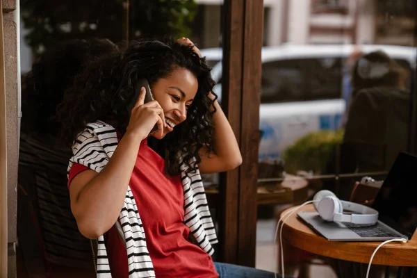 年轻的黑人妇女坐在外面的咖啡馆里 一边笑一边用手机说话 — 图库照片