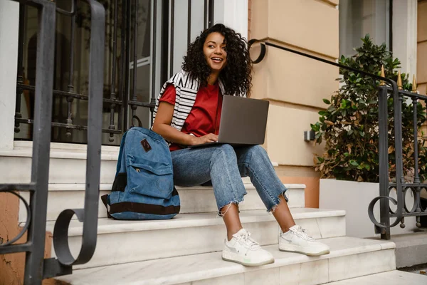 年轻的黑人妇女坐在楼梯上 在室外建筑时与笔记本电脑一起工作 — 图库照片