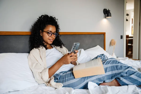 stock image Black young woman in eyeglasses using mobile and reading book on bed at home