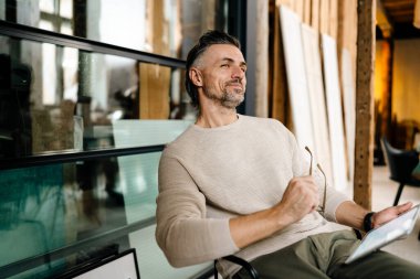 Orta yaşlı beyaz bir adam gülümsüyor ve elinde tablet bilgisayarla modern ofiste oturuyor.