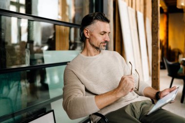 Modern ofiste kapalı otururken tablet bilgisayar kullanan orta yaşlı beyaz bir adam.